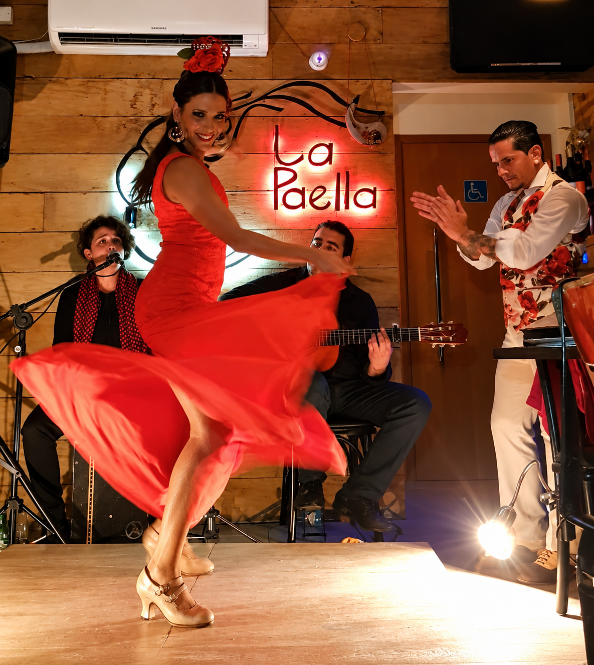 Bailaora de vestido vermelho com volantes em meio a um giro, em salão de paredes de tijolos com casal à mesa