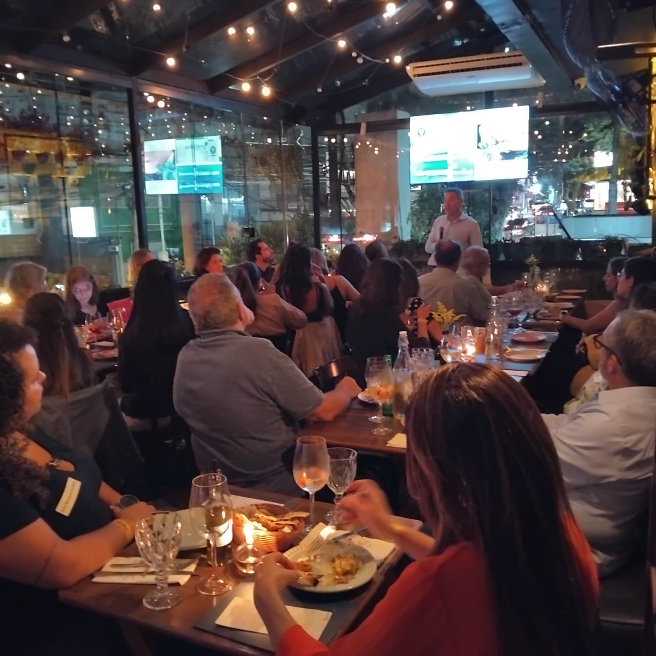 Convidados jantando no deck envidraçado do La Paella à noite, com apresentação ao fundo