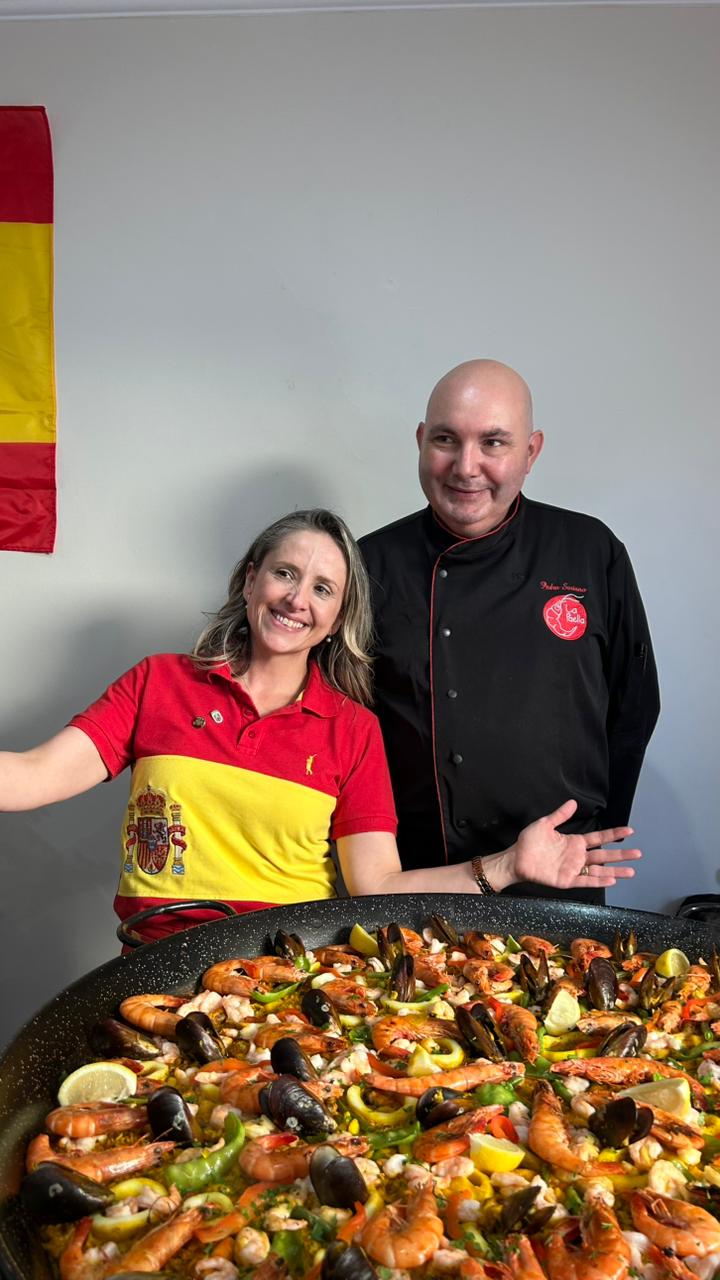 Chef Pedro Soriano ao lado de cliente diante da paella gigante, com a bandeira da Espanha ao fundo