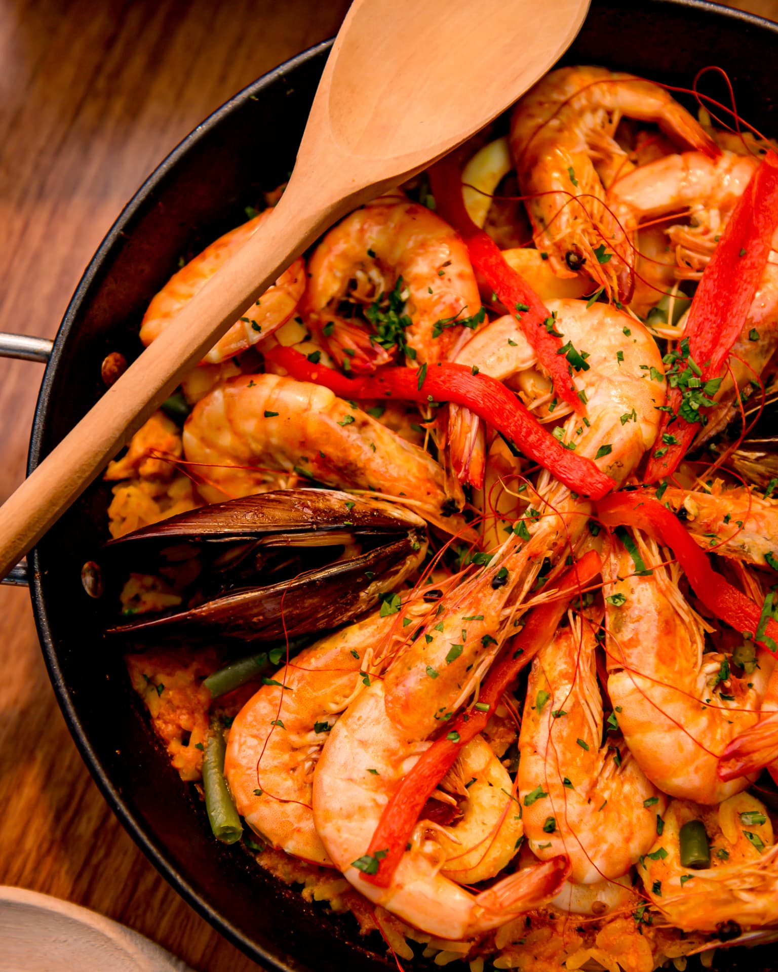 Paella de frutos do mar na paellera com camarões, tiras de pimentão vermelho, mexilhão e ervas, atravessada por uma colher de pau